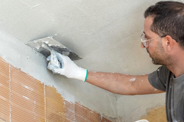Craftsman spreading plaster with metal float over a ceiling. Men in dirty gloves and gray shirt with dust and plaster on work clothing during construction or renovation process
